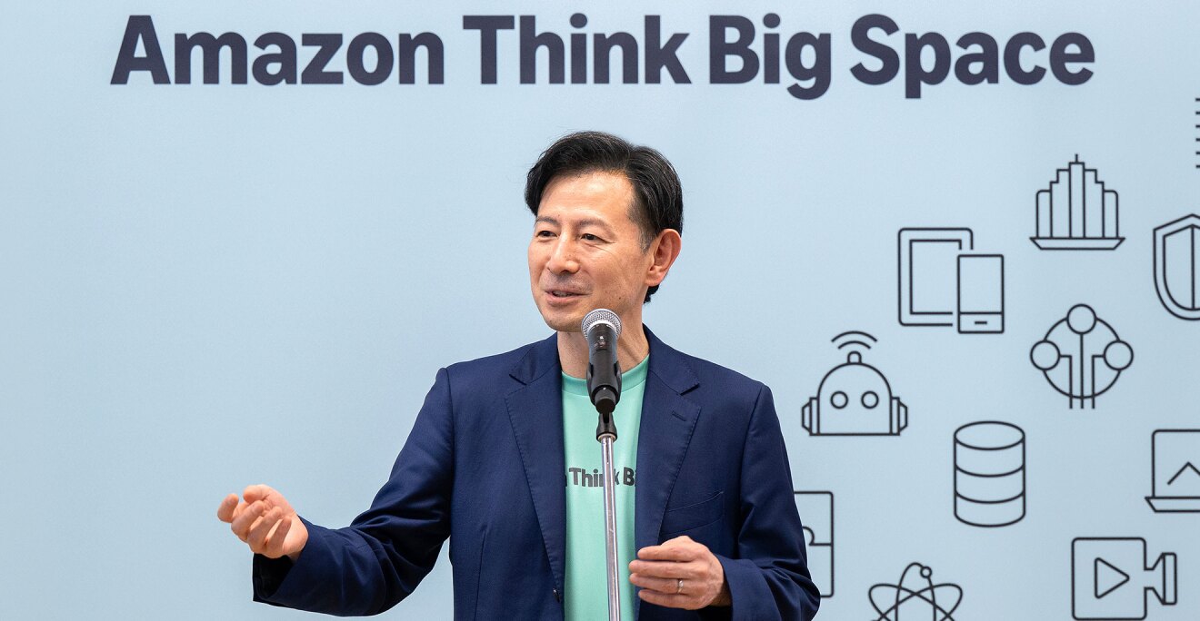 Amazon Think Big Spaceと書かれた青い背景の前に立つ人物。ミントグリーンのTシャツに紺のジャケットを羽織り、スタンドマイクに向かってスピーチしている。背景の右側にはデバイスやロボット、セキュリティのシールドなどを表す線画アイコンが多数配置されている。