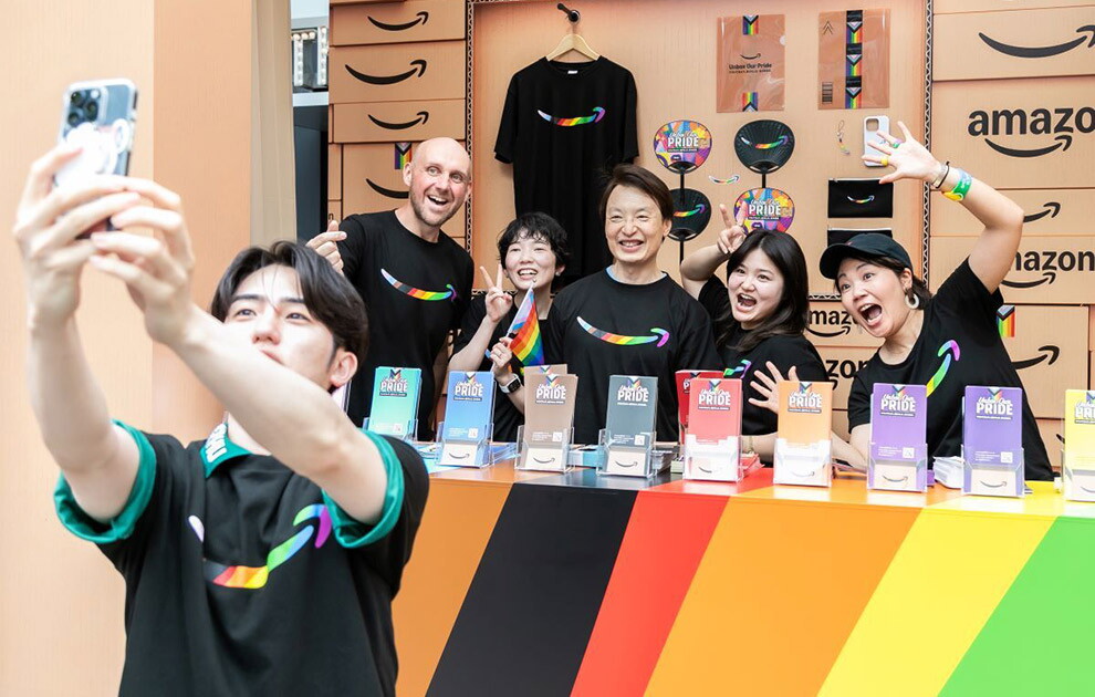 段ボールに囲まれたブース内で黒いTシャツを着た5人がポーズをとっている。その手前にスマートフォンで自撮りをしている男性