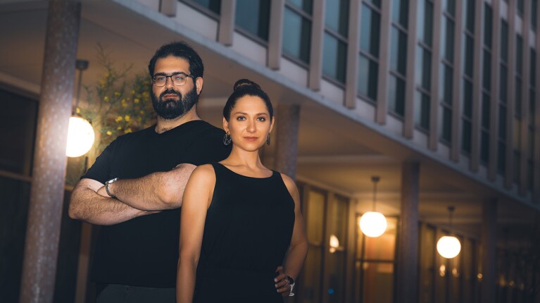 A photo of a man standing with his arms crossed next to a woman whose hand is on her hip. They are standing outside.