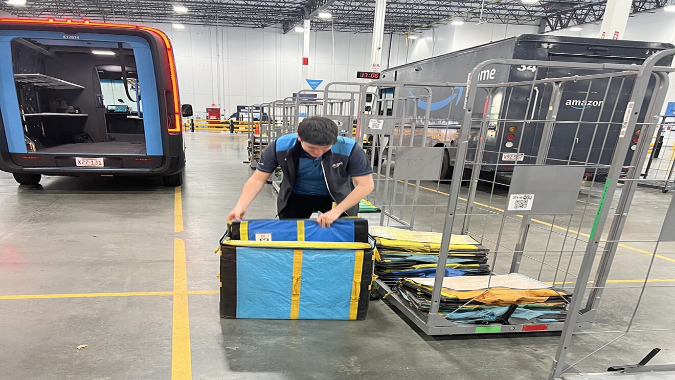 An Amazon delivery driver unloads totes from a Rivian van into a fulfillment center.