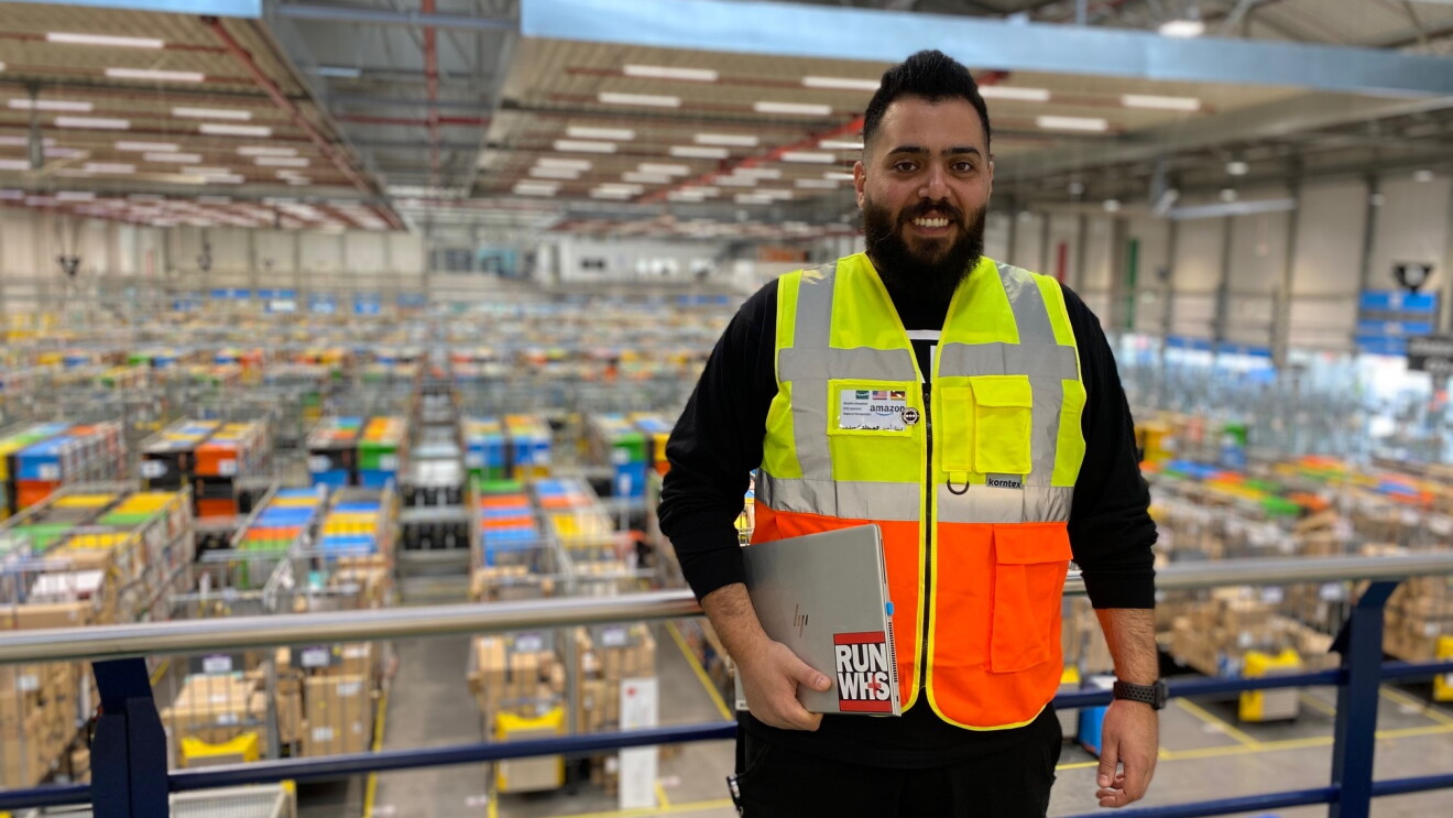 Spezialist für Arbeitssicherheit bei Amazon Mustafa Almujahed steht mit einer gelb-orangenen Weste bekleidet samt Laptop in einem Logistikzentrum.
