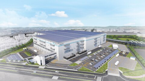 大規模物流施設の空撮イメージ。広大な屋根全面に太陽光パネルを配置。白い建物の側面に「amazon」の文字。周囲には駐車場、ソーラーカーポート、緑地帯、道路が整備され、建物前には大型トラック用の荷捌きエリア。背景には市街地と遠くに山々が広がる。