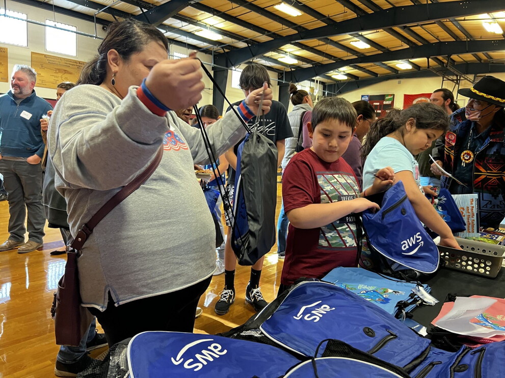 An image of a group of people picking up backpacks from a table. The backpacks have the AWS logo on them.