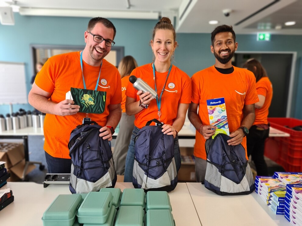 Amazon unterstützt sozial benachteiligte Schüler beim Schulstart. Amazon Mitarbeiter:innen stehen an einem Packtisch und zeigen den Inhalt der Schulrucksäcke.
