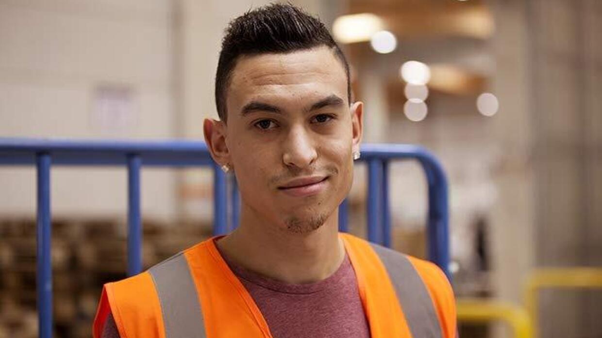 Un employé d'un centre de distribution d'Amzon en France sourit dans un décor de centre logistique. Il porte un gilet de sécurité.