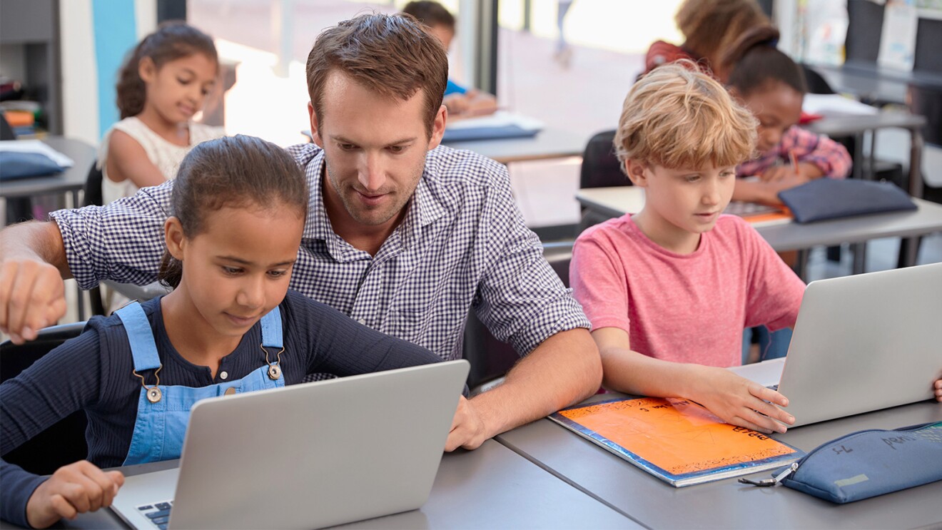 An image of a classroom of kids learning. There is a teacher showing one student something on their laptop.