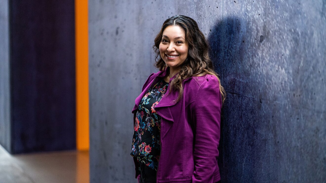 An image of a woman smiling for a photo standing in front of a dark gray and blue wall at an Amazon office building.