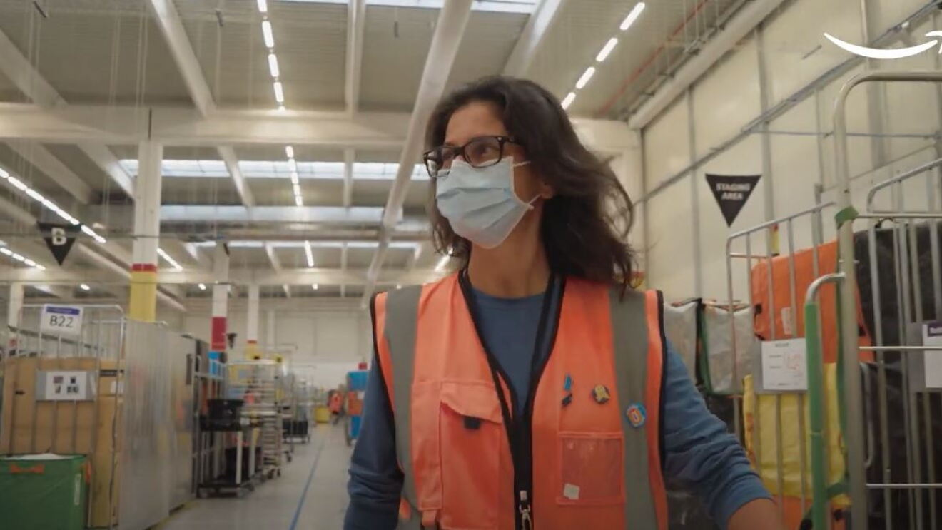 Mezzo busto di una donna che cammina in un centro logistico Amazon. Indossa un giubbino di sicurezza arancione e una mascherina chirurgica sul volto. 