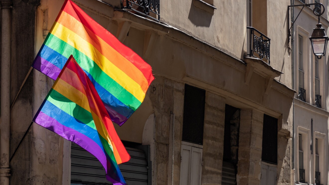 Two LGBTQ flags blow in the wind while attached to the side of an old historical building.