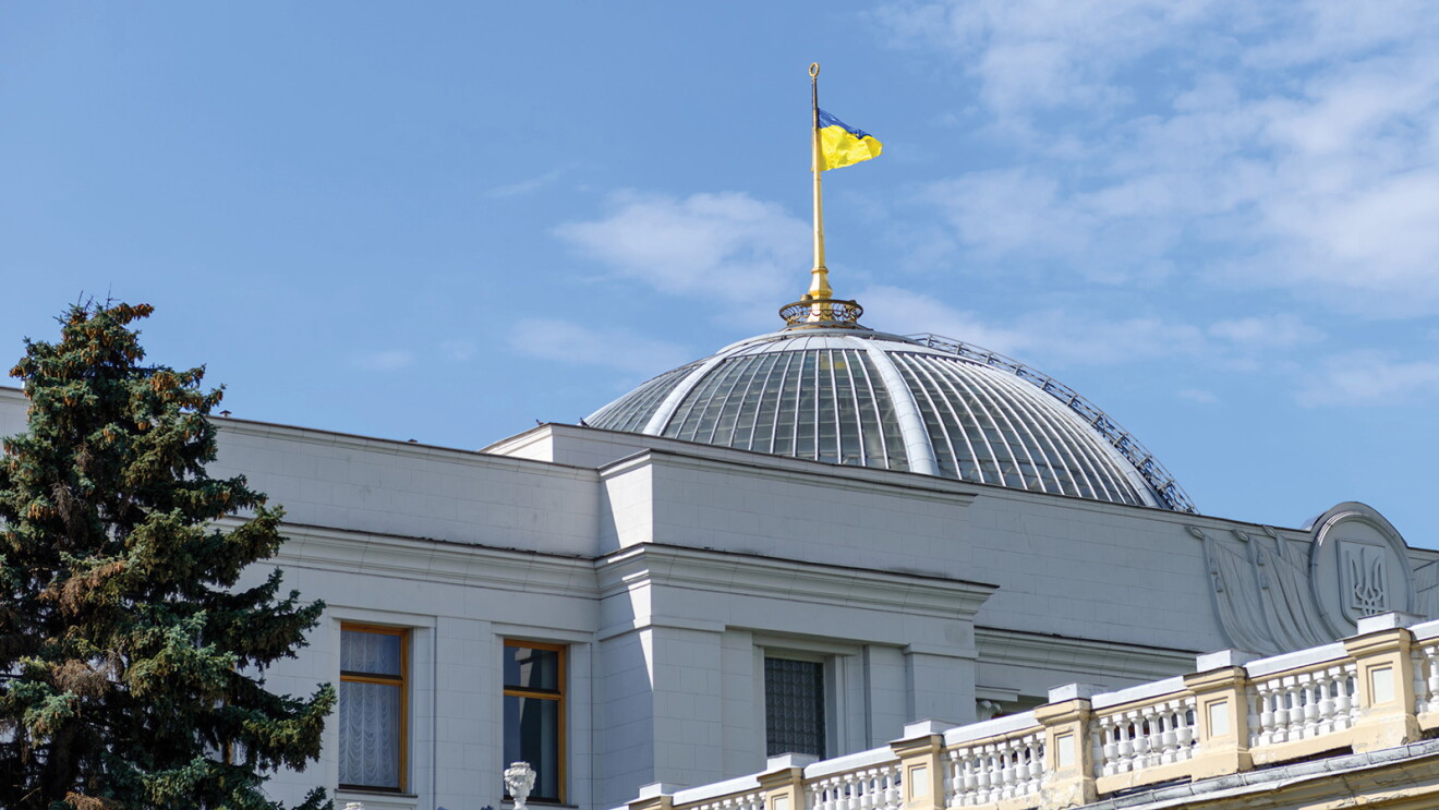 An image of the parliament building in Ukraine. There is the Ukraine flag flying atop the building and a blue sky in the background.