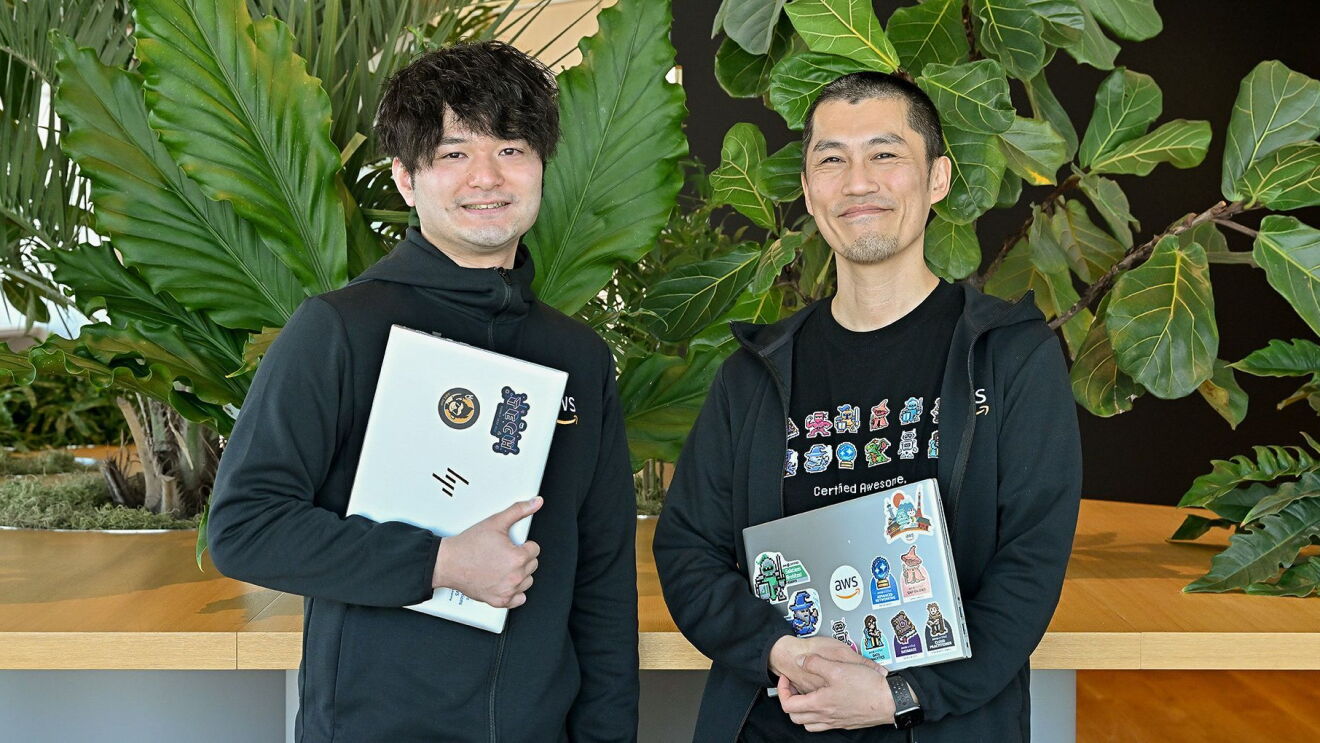 黒いシャツを着ている2人の男性がパソコンを持ちながら笑っている