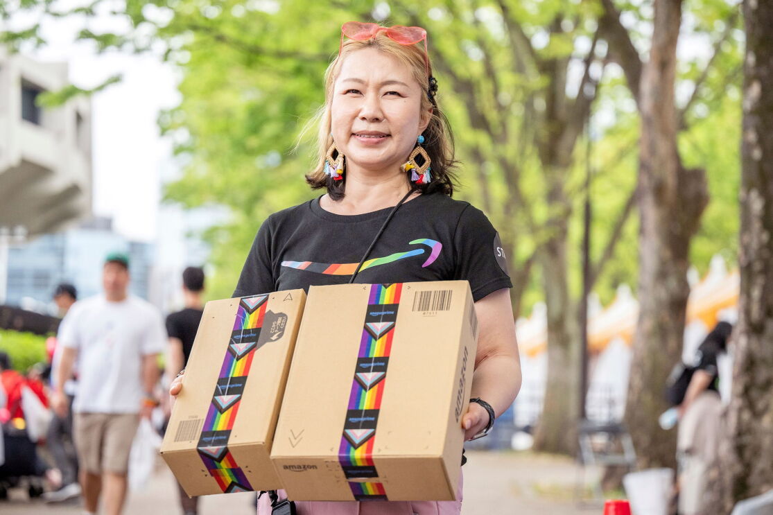 Amazonのロゴが入ったTシャツを着て、赤いサングラスを頭の上に載せている女性。手にはレインボーテープが貼られた段ボール箱を持っている