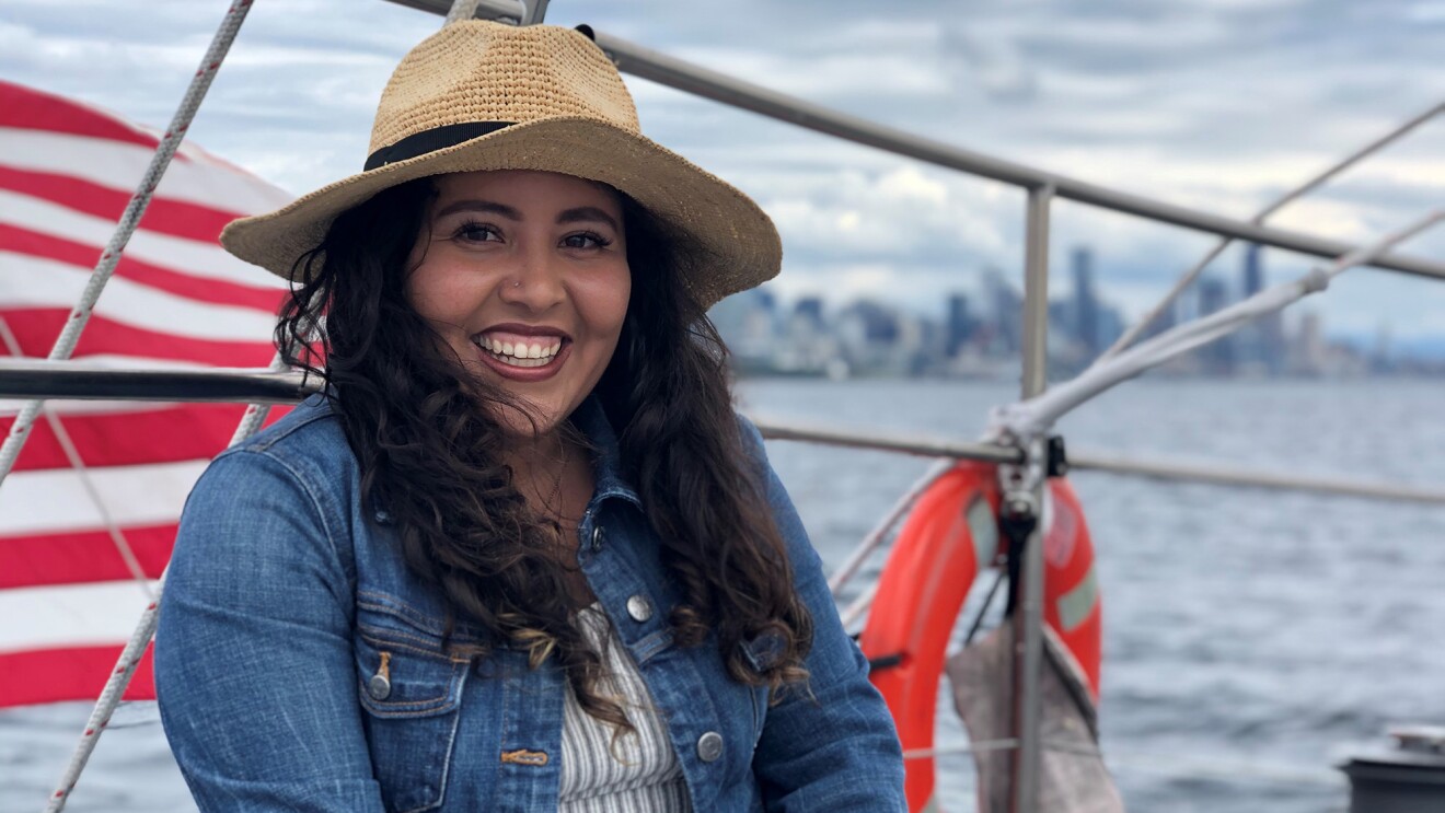 An image of a woman smiling for a photo while sitting on a sail boat. The American flag flies behind her on the boat.