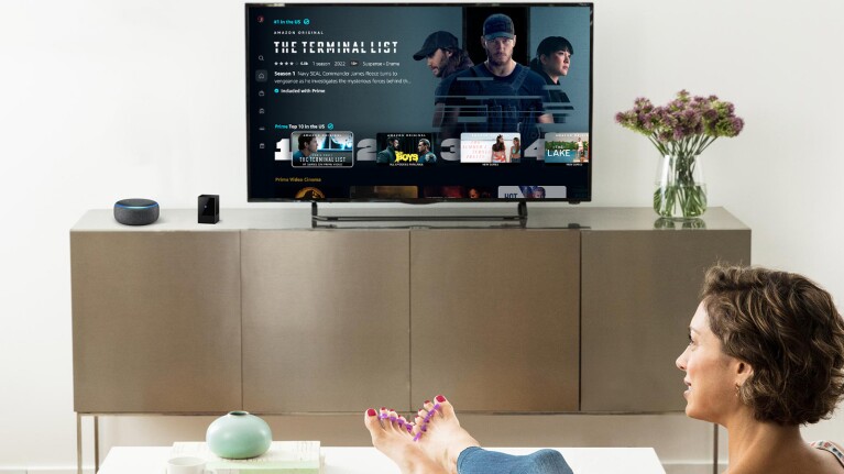 An image of a woman sitting in a living room watching Prime Video. There is an Amazon Echo device next to the TV.