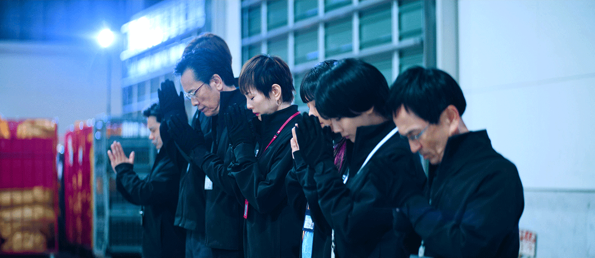 赤や緑の、ケージ状の台車が並ぶガレージ内で、そろいの黒い作業服を着た7人が横一列に並ぶ。全員、目を閉じて顔の前で合掌している。