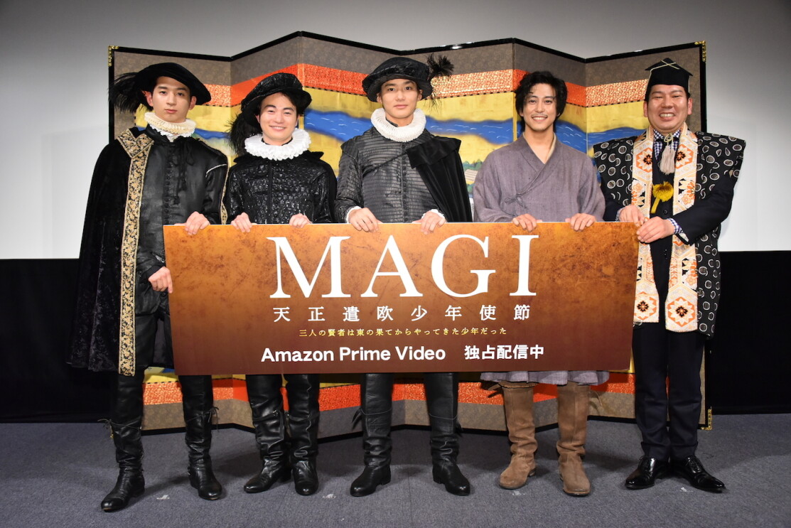 極彩色の屏風を背に、西洋の歴史衣装や着物を着て並ぶ5人の男性。全員で茶色のボードを掲げ持っている。