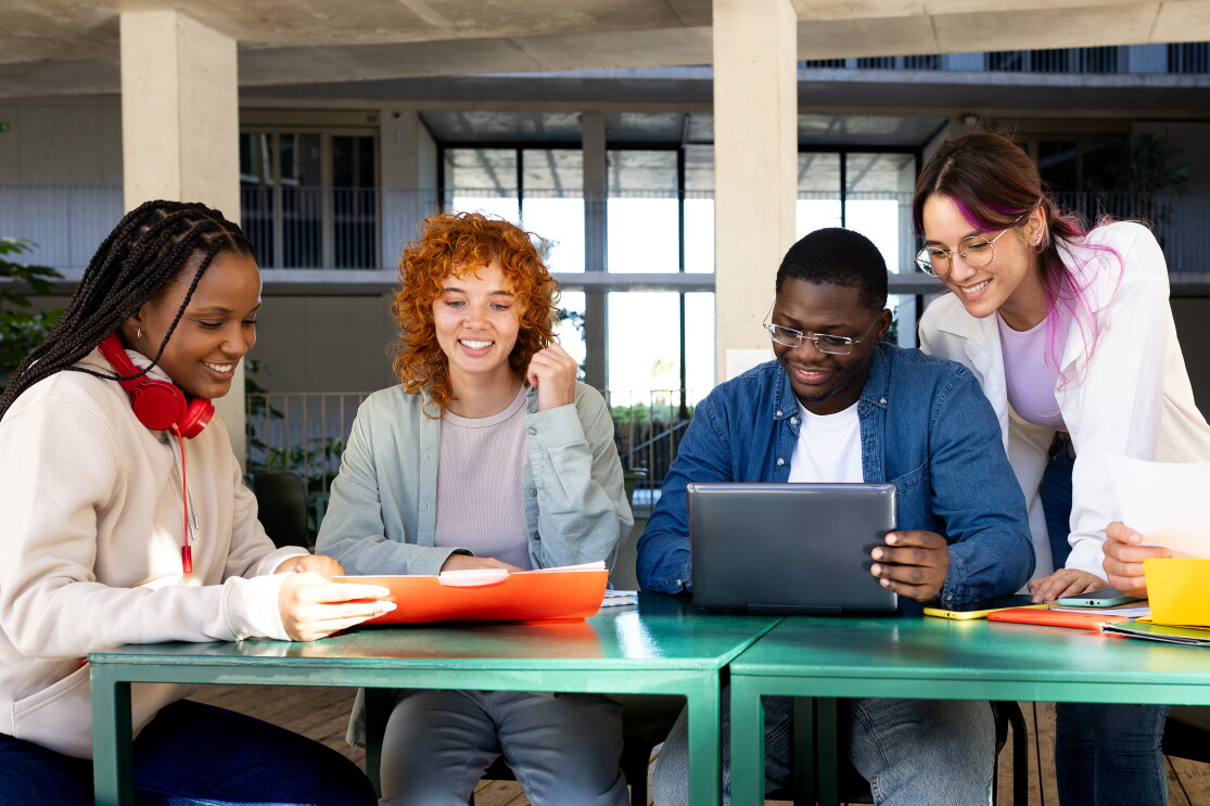 Four people sitting at a table looking at books and a computer screen. 