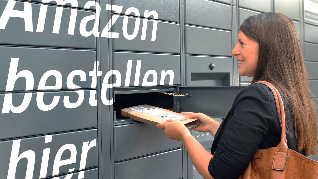 Der Bildausschnitt zeigt graue Paketschließfächer und den weißen Schriftzug "Amazon bestellen hier ...". Rechts ist eine junge Frau mit langen braunen Haaren zu sehen, die ein längliches, flaches Amazon Paket aus dem Schließfach entnimmt.