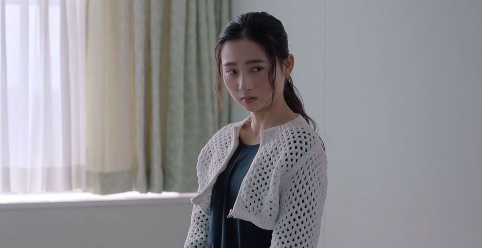 白い壁の室内に立つ人物のバストアップ。白く丈の短い上着と中に紺色の服を着用、右側を横目で見ている。人物の左側にレースのカーテンと薄い緑色のカーテン。