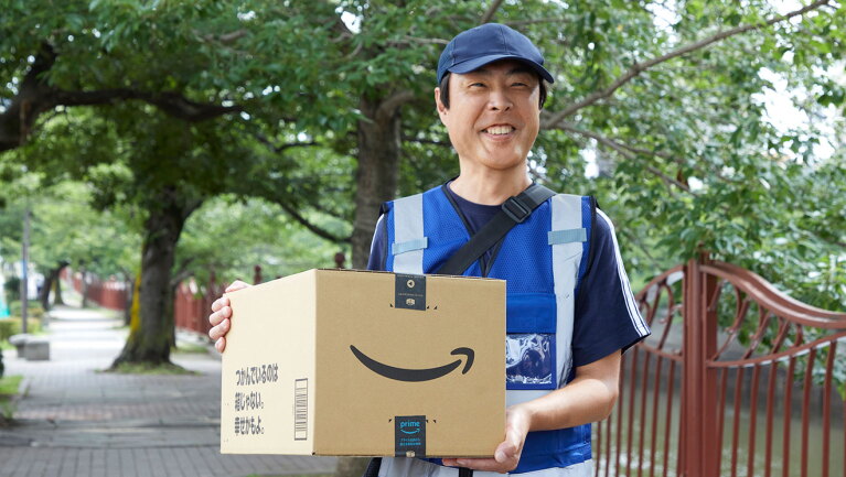 緑の葉が豊かに茂った街路樹を背に、ダンボール箱を抱える笑顔の配送スタッフ。右側には川が流れている。