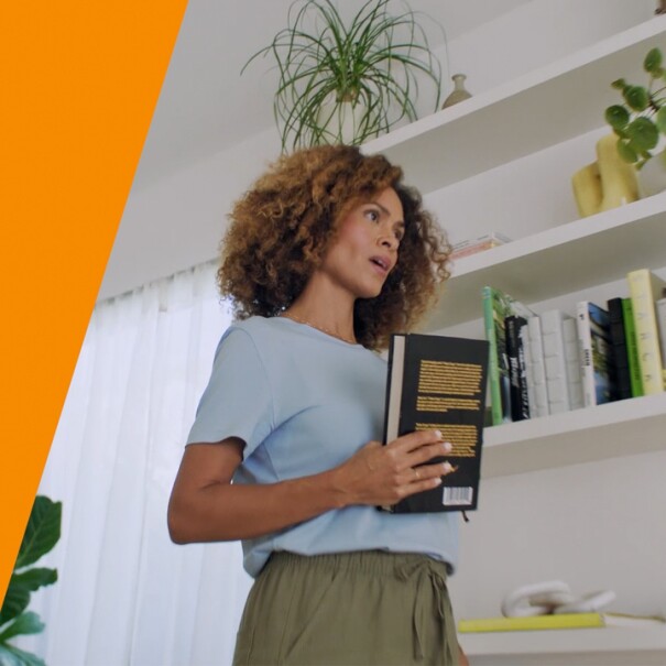 An image of a woman holding a book while talking to Alexa. There is an orange stripe on the left side. 