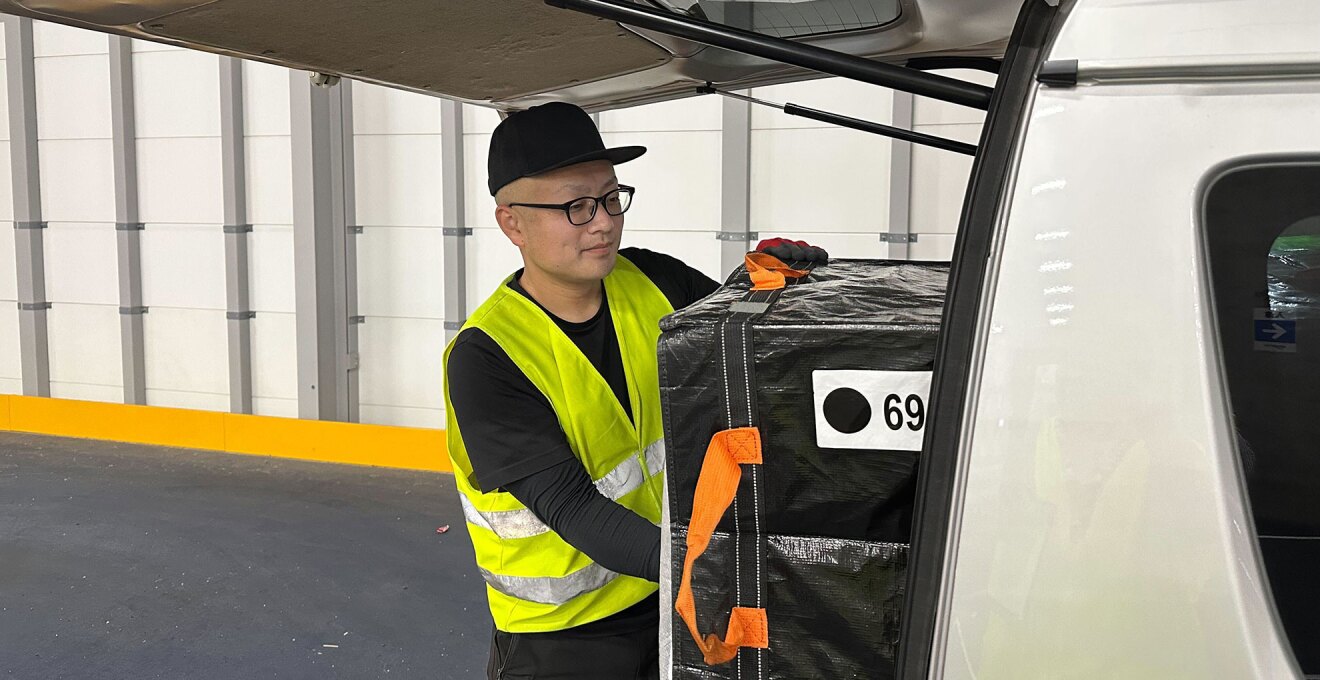 建物の外で、跳ね上げ式のバックドアから車に大きな荷物を積み込む男性。黒い帽子とシャツ、蛍光イエローのベストを着用している。荷物は黒で、オレンジ色の持ち手つき。