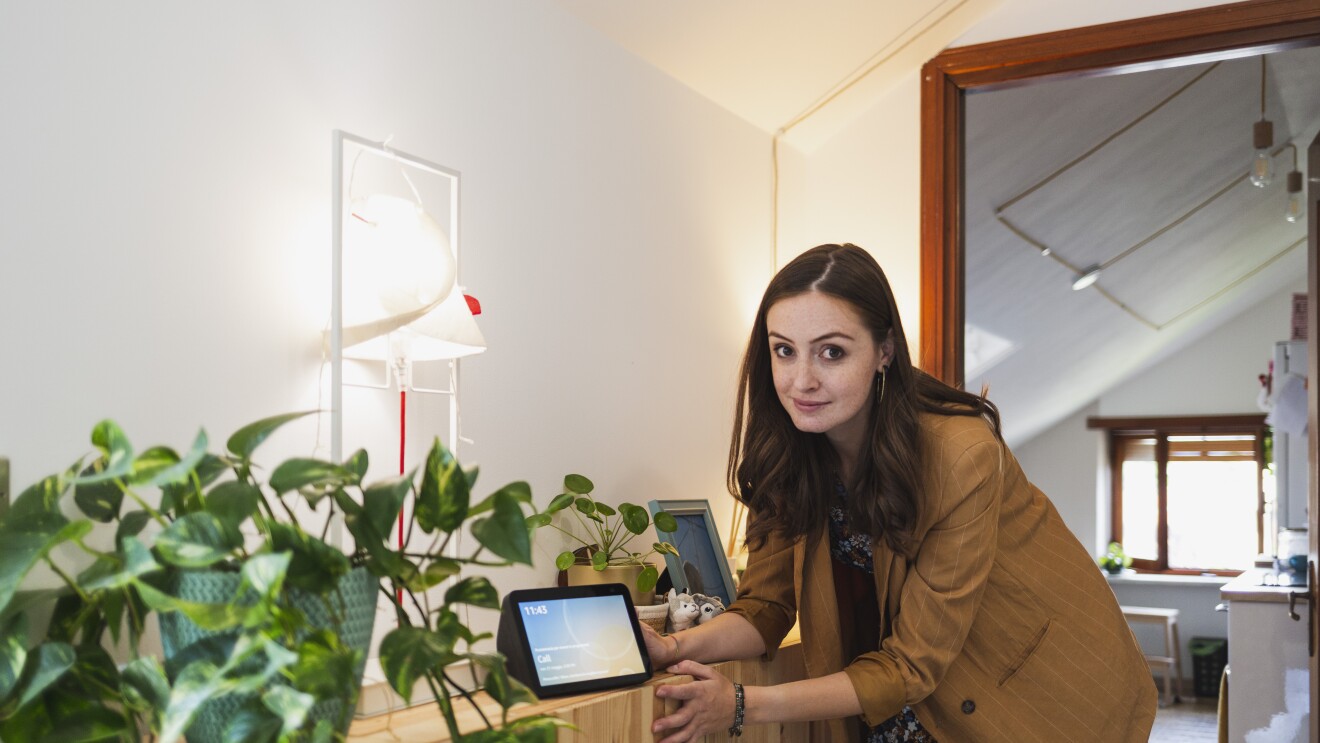 Elisa Reale, al centro della foto, è chinata verso un mobile. Guarda in camera, vicino alla mano destra ha un dispositivo Echo Show con lo schermo acceso. In primo piano, posata sullo stesso mobile, si vede una pianta in un vaso verde. Elisa indossa una giacca beige a righe bianche, ha i capelli lunghi e mossi. Sotto la giacca indossa una gonna a fiori colorata. 