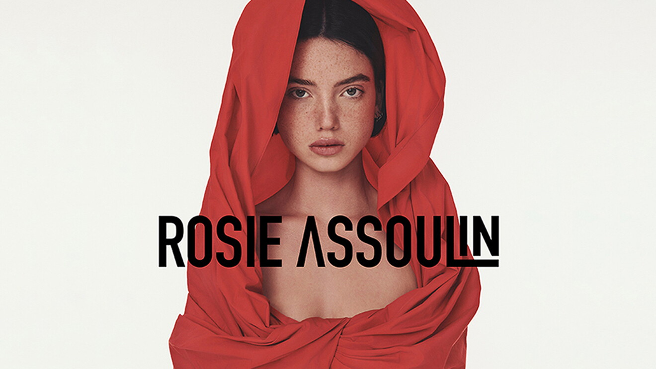 A woman wearing a red hoodie piece of clothing. The image has text overlay that reads, "Rosie Assoulin."