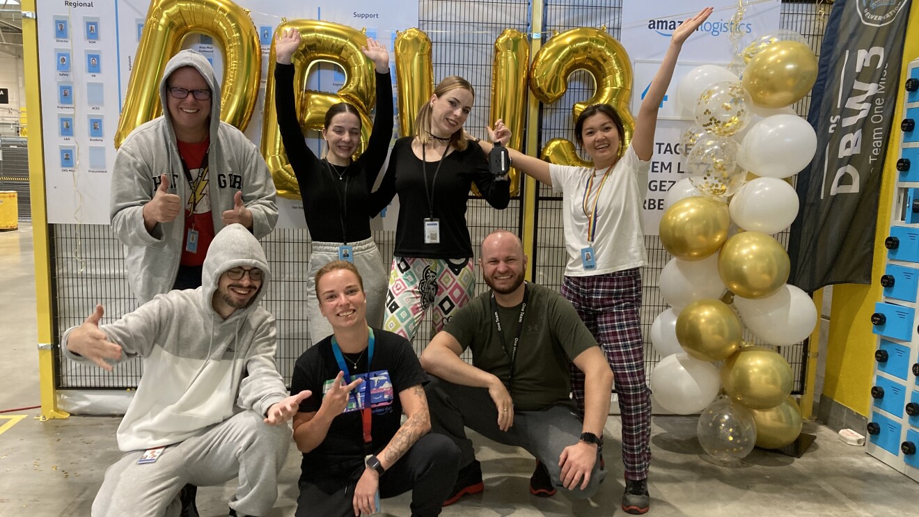 Mitarbeitende aus ganz Baden-Württemberg nahmen an der Initiative „Amazon Goes Gold for Kids with Cancer“ teil und posierten vor Wand mit weißen und goldenen Luftballons.