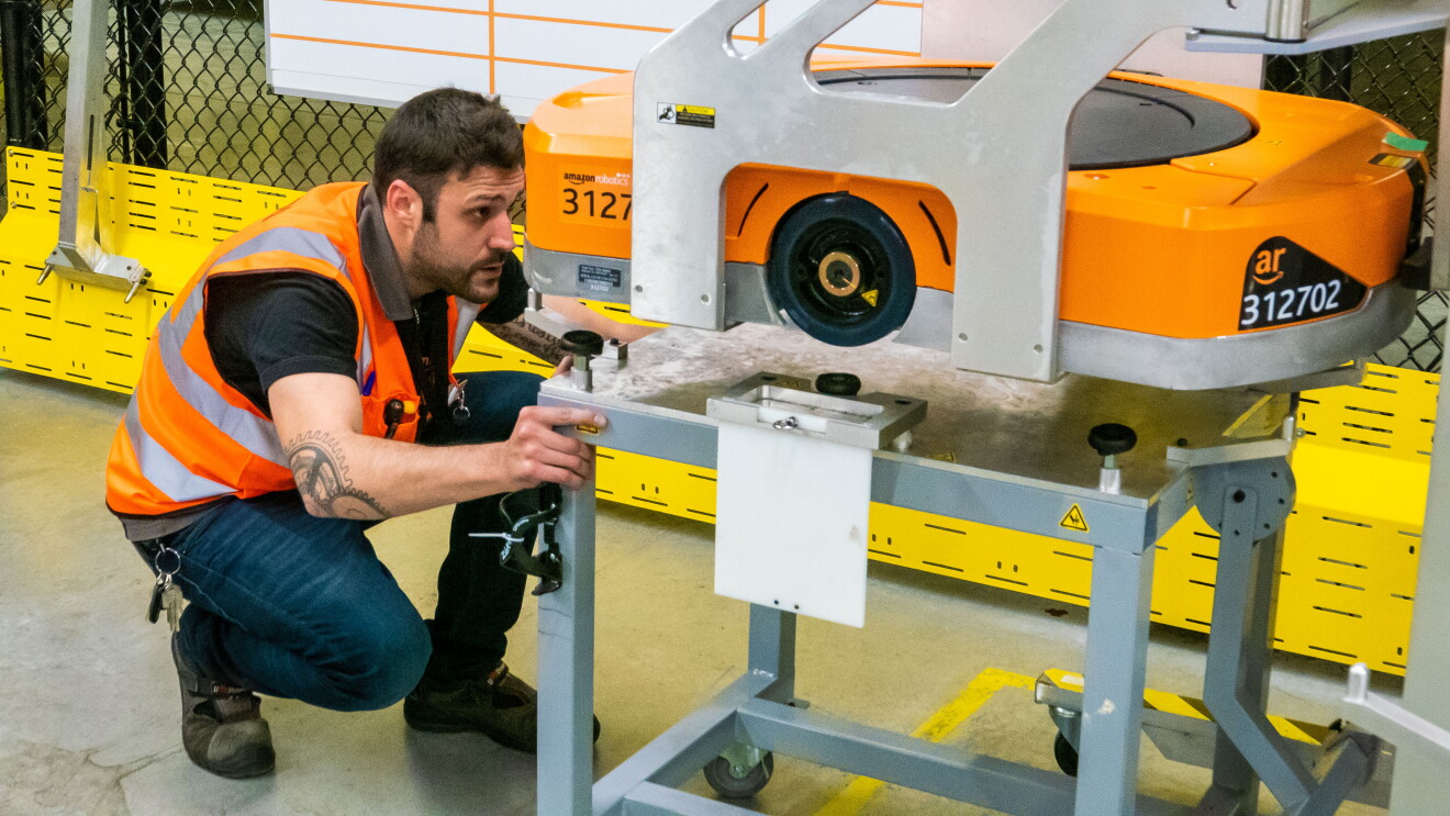 Raúl Cruz, técnico senior de mantenimiento en robótica, reparando un robot. En primer plano está Raúl, con el pelo negro y barba, vestido con una camiseta negra y el chaleco naraja. Observa atentamente un robot que está en una mesa pero alzado por una grúa, con lo que Raúl puede observar la parte inferior. 