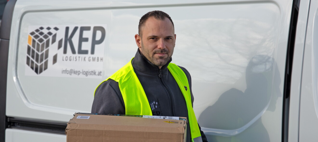 Ein Mann mit Bart in gelber Sicherheitsweste steht vor einem weißen Liefer-Van mit Aufdruck KEP Logistik GmbH. Er hält ein Paket in der Hand.