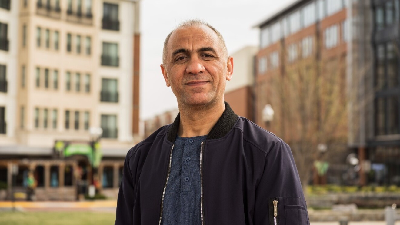 Khalid Noorzad stands outside in a park between buildings and grins at the camera.
