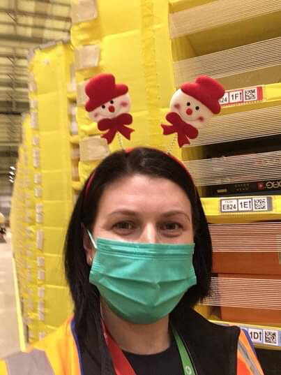 Diana Campean, Associate at Amazon's fulfilment centre in Dunstable pictured in front of lots of yellow pods full of inventory. Diana is wearing a green face mask and a festive headband with two snowmen.