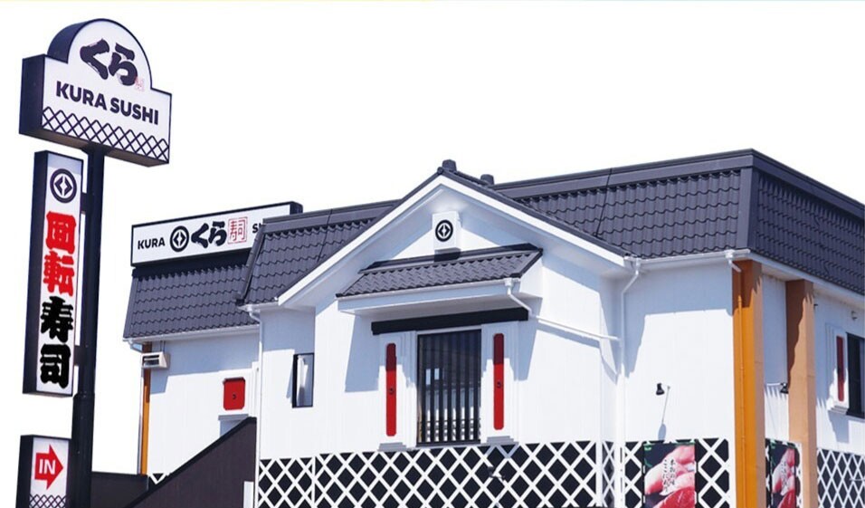 白背景の、回転寿司店舗の外観イラスト。白壁とグレーの瓦の和風の建物で、壁の下側は黒字に白の格子模様。左側に「くら」「回転寿司」の高い看板が立っている。