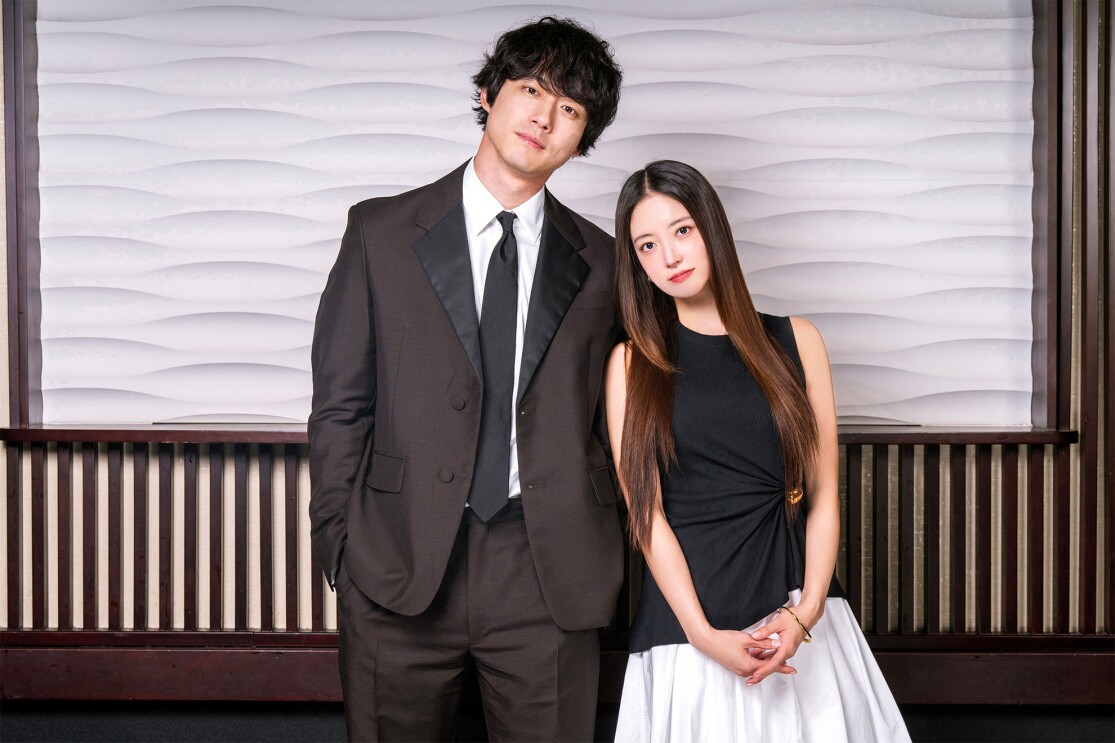 ウェーブヘアの男性がスーツを着ており、ロングヘアの女性が黒いトップスを着て、お互いに寄り添っている。