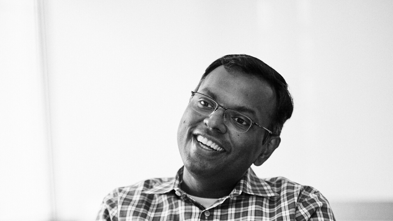 A man wearing a plaid shirt and glasses sits at a table in a conference room, explaining something to the photographer. 