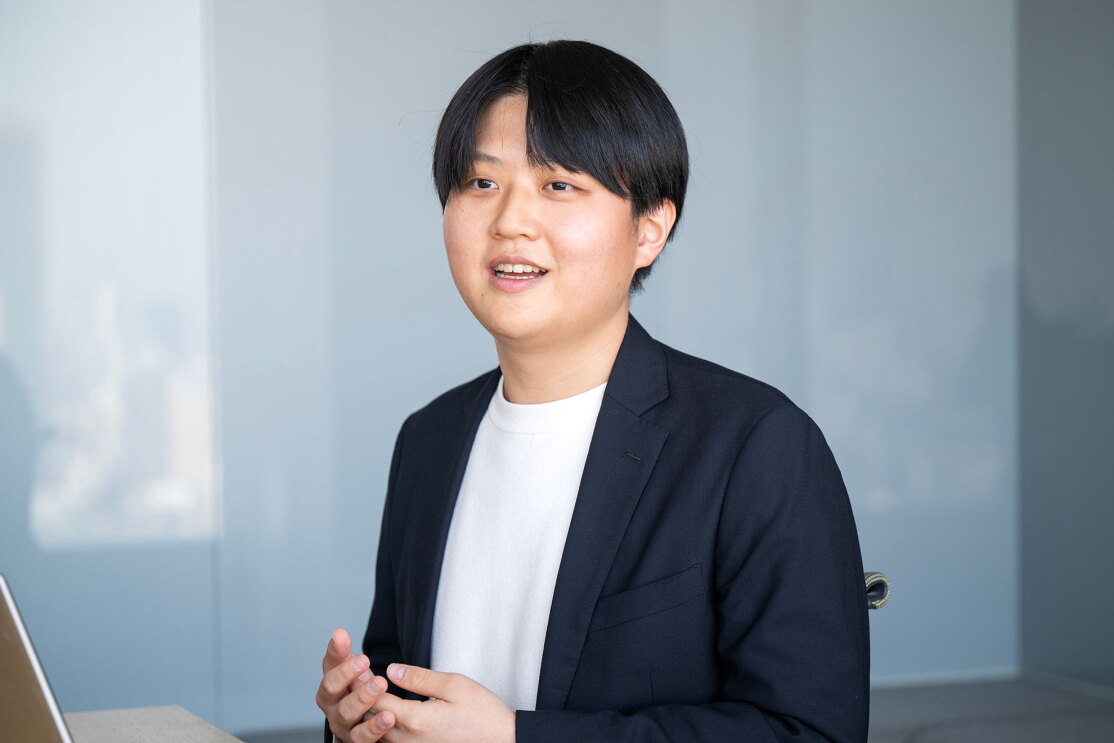 グレーの壁を背景に、ネイビージャケットと白のトップスを着用した男性が着席している。両手のジェスチャーを交えながら会話をしている。