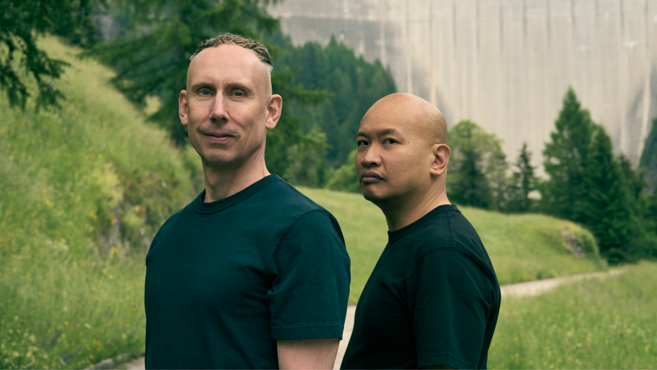 Two men dressed in black looking seriously into the camera in a green, hilly landscape