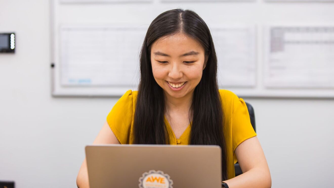 An image of a woman wearing a yellow shirt seated in front of a laptop in an office setting.