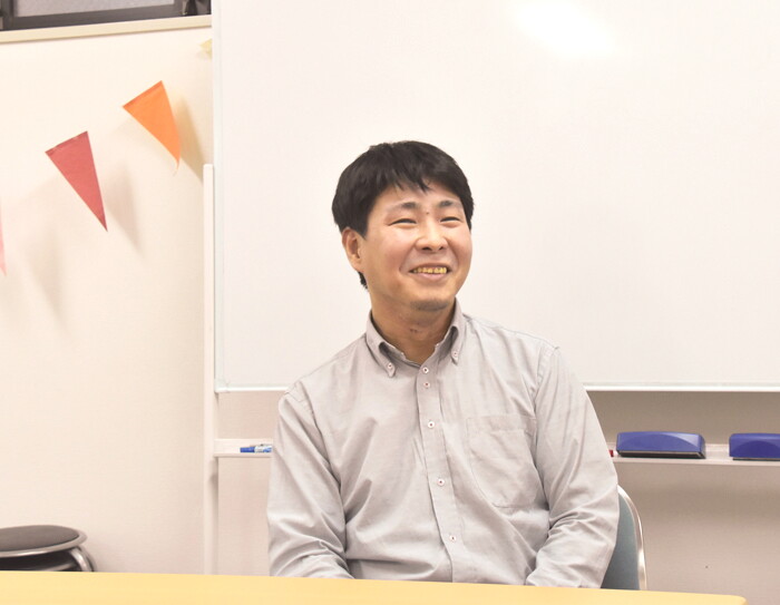 白い壁、ホワイトボードを背に着席して話す男性。薄いグレーのシャツ。左側に赤とオレンジのガーランド。