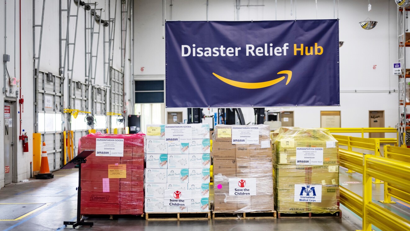 An image of the inside of an Amazon fulfillment center. There are boxes lined up with clear wrapping on them, and they are labeled with different charity names as donations. There is a large banner hanging from the ceiling above the boxes that says "Disaster Relief Hub" with the orange Amazon logo below it.