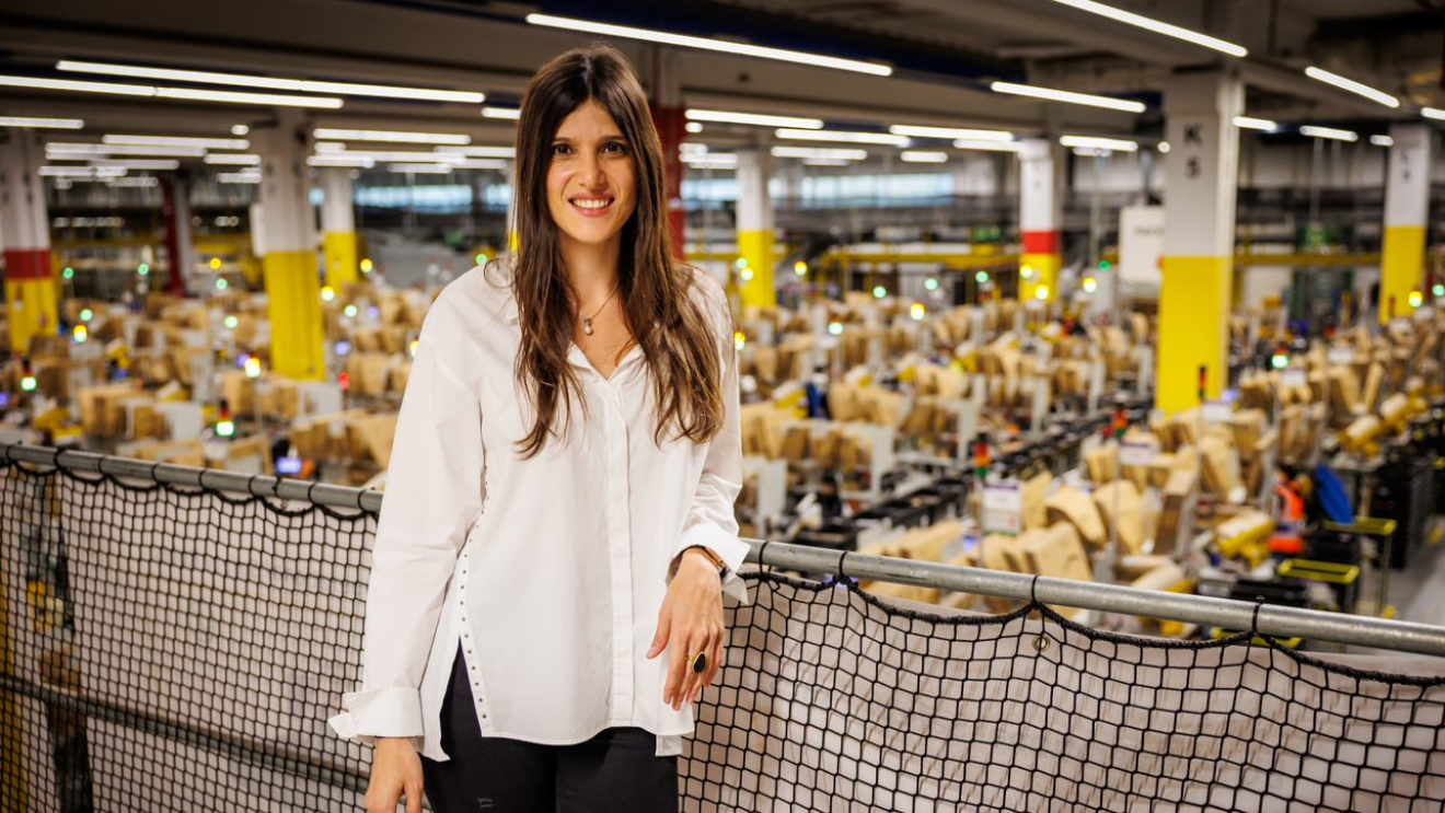 Geraldine Leiros, directora general del centro logístico robotizado que Amazon tiene en Illescas (Toledo). Dentro de un centro logístico aparece del fondo desenfocado con estantería de paquete. Geraldine aparece en un balcón con una red que protege la barandilla de color negro. Ella se encuentra de pie, con una camisa blanca por el muslo y unos pantalones negros. Lleva el pelo largo castaño a la altura del pecho, con un anillo negro grande en la mano derecha y un collar. 