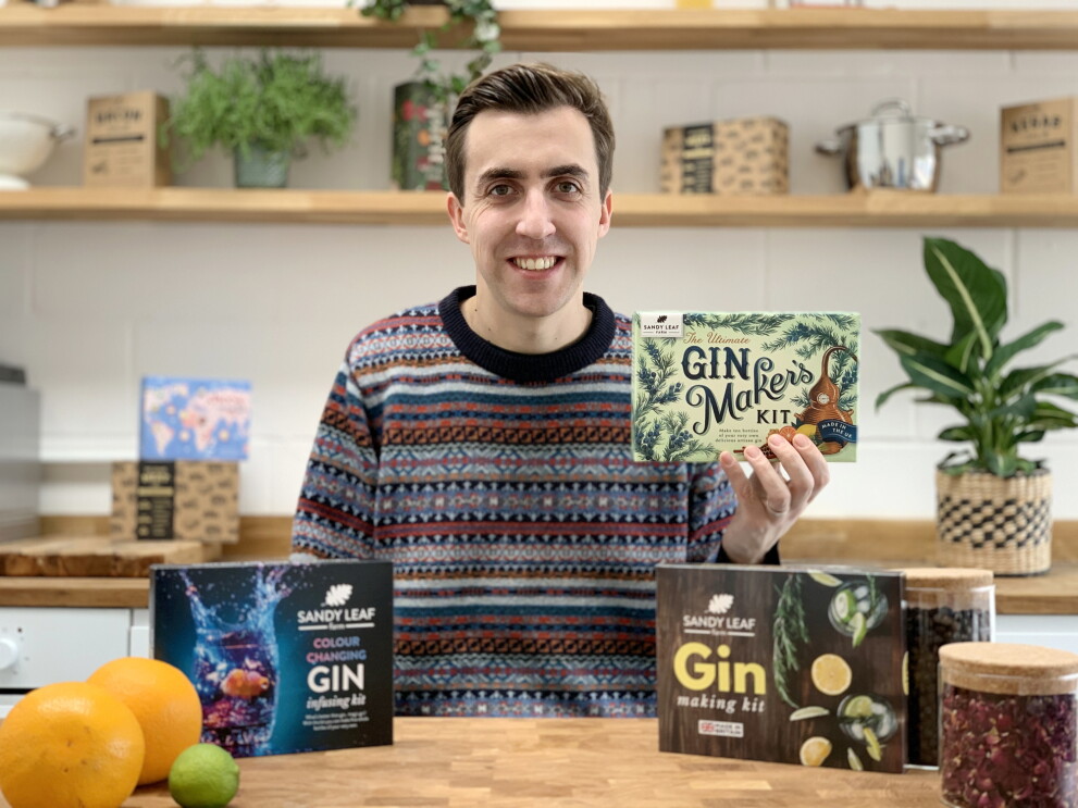 Scott MacDonald from Sandy Leaf Farm at a table with the gin making kits in front of him. He is also holding a box containing the products.
