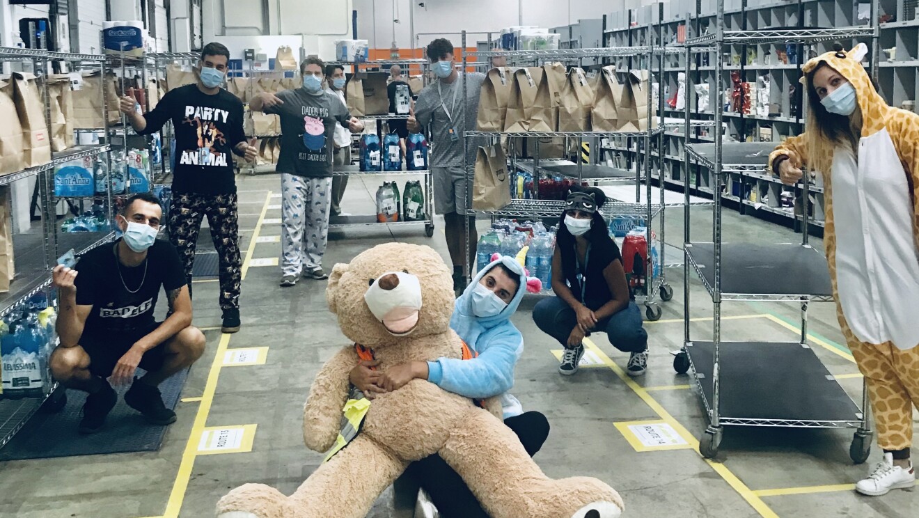 Dipendente Amazon posa con un peluche a forma di orso in un centro logistico. Dietro di lei, altri dipendenti  indossano pigiami.