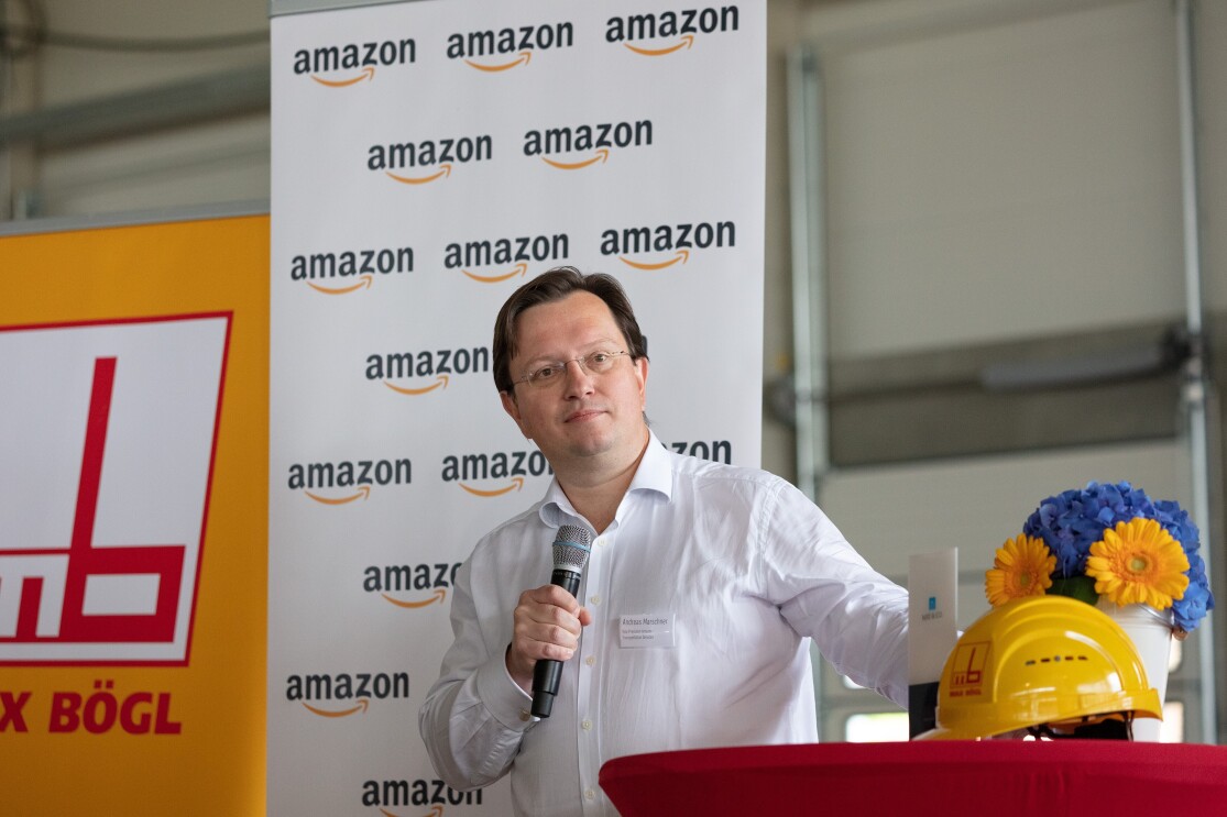 Ein Mann vor einem rot überzogener Stehtisch. Im Hintergrund sind Rollups mit Audruck amazon und Bögl zu sehen. Auf dem Tisch stehen ein gelber Bausstellenhelm sowie gelb-blaue Blumen