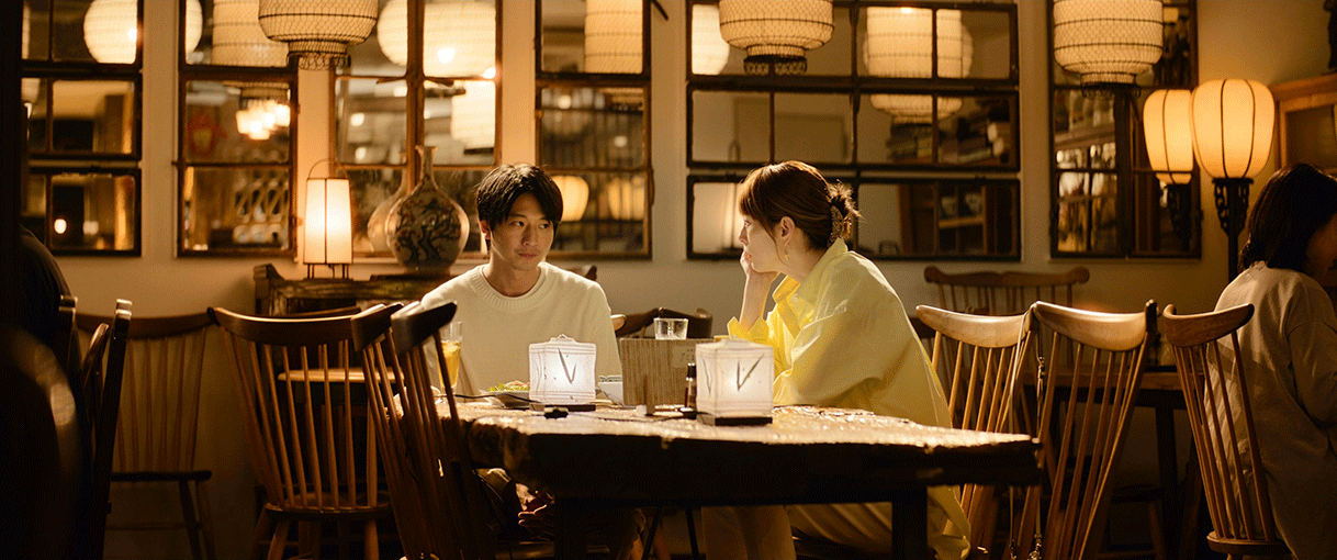 落ち着いた色調のインテリア、多数のランタンが灯るレストラン店内。テーブルにL字座りする2人の人物。右側の女性が片手で頬杖をつき、左側の男性と見つめ合う