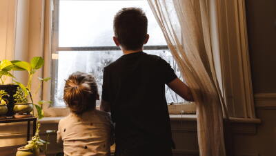Doa heramos: una hermana de unos 3 años y un hermano de unos 7 años. Los dos están de espaldas y mirando por una ventana. El niño va con una camiseta negra de manga corta. La niña, con una camiseta blanca y con lágrimas de colores. Fuera de la ventana se ve todo blanco y parece que hay un árbol con nieve. El niño con la mano derecha aguanta unas cortinas blancas. El marco d e la ventana es blanco y de madera. En laparte izquierda de la foto hay un mueble con 4 plantes y se nota que entra la luz de una lámpara. 