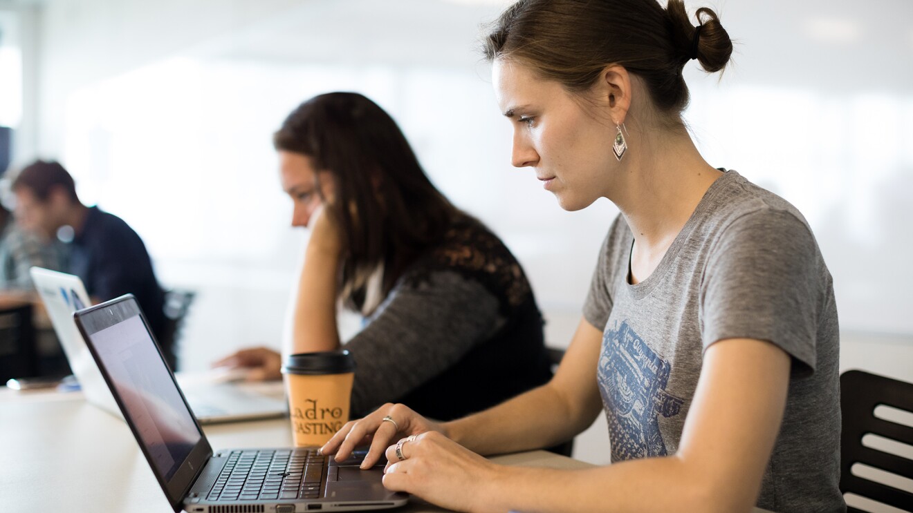 A woman in a t-shirt works on a laptop computer in a workplace setting. Next to her, another woman works on a laptop, and behind them, men sit at a table.