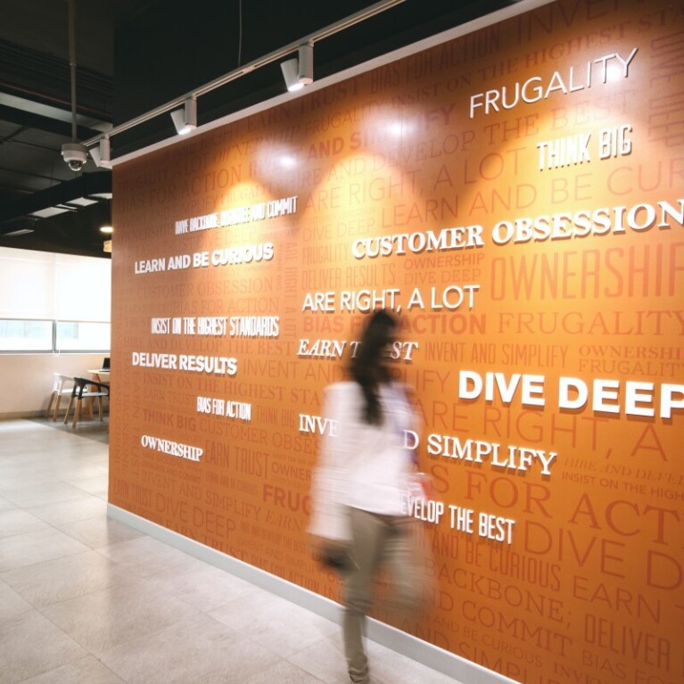 Amazon's Leadership Principles featured on a wall in an Amazon office building. A woman is walking in front of them.