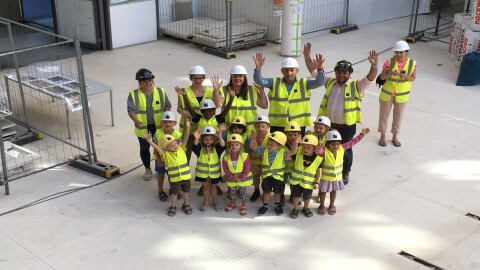 Pupils between age 4 and 6 wave at the camera with Amazonians and their teachers, all wearing neon yellow working vests and construction helmets.  They are at the construction site of Amazon's new headquarters building in Luxembourg.